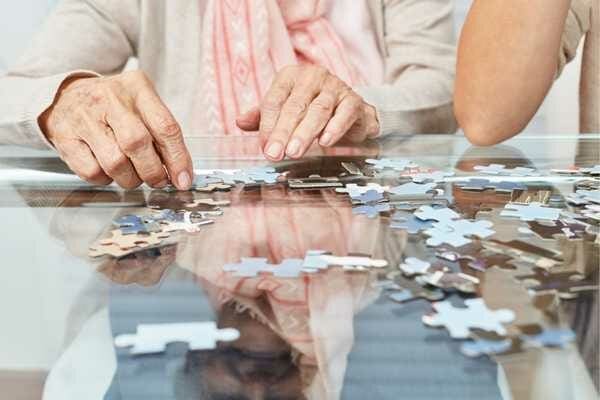 A pair of hands belonging to a senior person is doing a jigsaw puzzle on a glass table